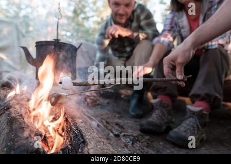 Gruppo di amici frittura dolce marshmallows su un fuoco, sopra la fiamma di cui un bollitore pende, in un campeggio, nella foresta. Foto Stock