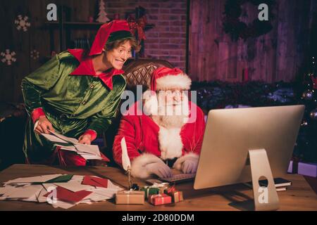 Ritratto fotografico di babbo natale ed elfo che guarda al computer schermo che tiene un mazzo di lettere Foto Stock