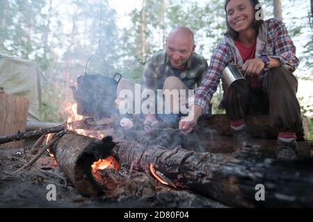 Amici felici friggono i marshmallows sopra un fuoco, sopra la fiamma di cui un bollitore pende, una donna con una tazza che aspetta una bevanda calda. Foto Stock