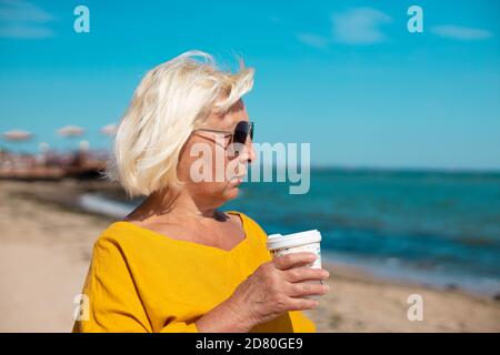 Caffè in movimento. Bella donna bionda che tiene la tazza di carta del caffè e che gode una passeggiata lungo la costa del mare Foto Stock