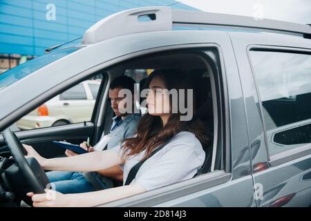 Giovane donna sconvolto prende l'esame di guida con istruttore di auto maschile serio. Due persone sedute in auto grigia con cinture di sicurezza allacciate. Concentrato giovani WO Foto Stock