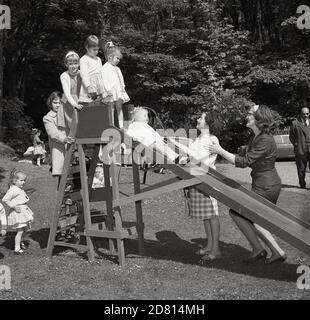 Anni '60, fuori su un campo in un parco, bambine in attesa del loro turno di scendere uno scivolo di legno, con le loro madri a portata di mano per catturarle, Fife, Scozia. Foto Stock