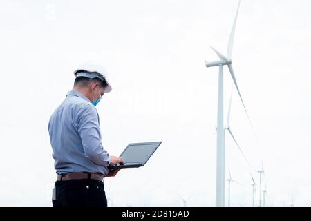 Ingegneri mulini a vento indossando maschera e lavorando su computer portatile con la turbina eolica in background Foto Stock