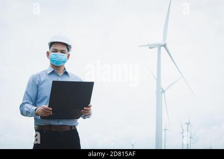 Ingegneri mulini a vento indossando maschera e lavorando su computer portatile con la turbina eolica in background Foto Stock