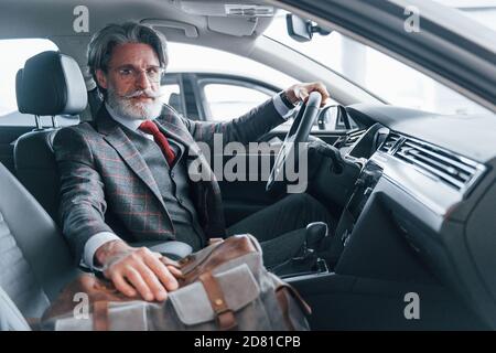 Uomo anziano alla moda con capelli grigi e baffi in vacanza con borsa all'interno dell'automobile moderna Foto Stock