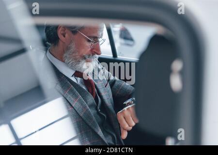 Uomo anziano moderno ed elegante con capelli grigi e baffi ai suoi orologi all'interno dell'automobile Foto Stock