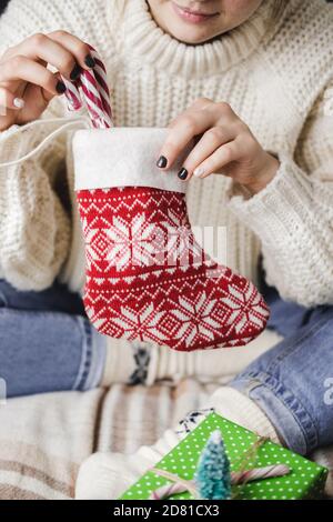 La giovane donna si siede sul plaid in felpa di lana lavorata a maglia accogliente e mette la canna lollipop a righe in calza di Natale per i regali. Preparazione per le vacanze invernali. WO Foto Stock