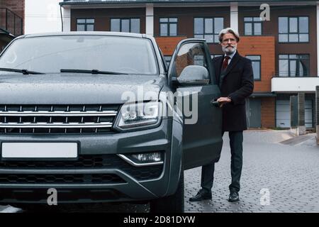 L'uomo anziano alla moda con i capelli e la barba grigi è all'aperto sulla strada apre la porta della sua auto Foto Stock