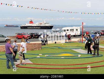 I turisti giocano a golf pazzo sul lungomare di Arran mentre il traghetto Caledonian MacBrayne arriva dalla terraferma, a Brodick Bay, Scozia, Regno Unito, Europa Foto Stock