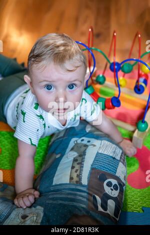 Cute bambino ragazzo strisciando sul letto di cuscino sdraiato sul pavimento dopo aver giocato con il gioco di labirinto di perle di legno e guardando sorpreso e curioso Foto Stock