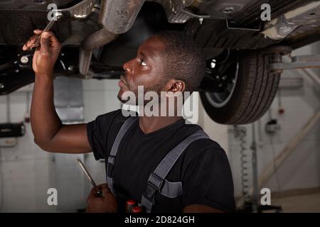 bel maschio africano che ripara il fondo dell'automobile, controlli ed esamini tutti i particolari. uomo duro in uniforme di lavoro Foto Stock