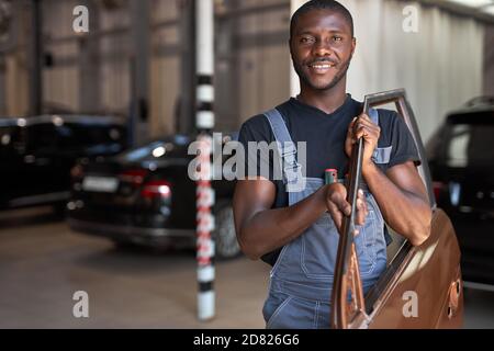 giovane meccanico africano al lavoro con parte separata della macchina, in posa alla macchina fotografica, in corso di riparazione Foto Stock