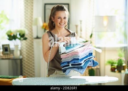 Ritratto di donna moderna sorridente in blusa di seta e pantaloni beige con ferro da stiro a vapore, asse da stiro e pila di abiti stirati piegati nella casa moderna Foto Stock