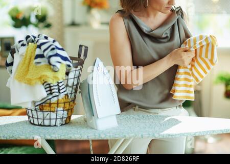 Closeup su donna in blusa di seta e pantaloni beige con ferro da stiro a vapore, cesto di vestiti lavati e asse da stiro infelice con lavanderia risultato in casa in s. Foto Stock