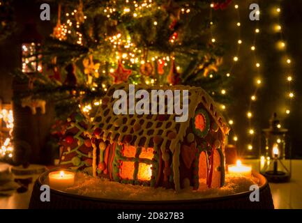 Casa di pan di zenzero colorata e decorata divertente con finestre di vetro caramello e candela che illuminano all'interno, bokeh albero di Natale sfocato. Foto Stock
