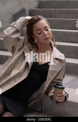 elegante e sottile bruna giovane in trincea alla moda e abito nero seduta su scala in cemento grigio con una tazza di caffè di carta marrone. Foto Stock