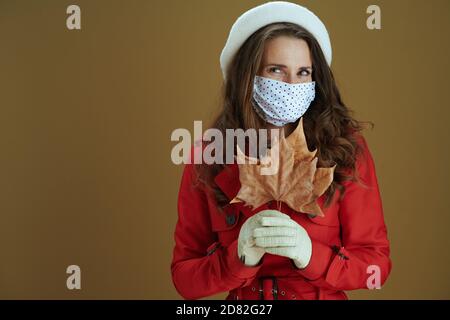 Vita durante la pandemia di covid-19. Ritratto di donna trendy pensiva in camice rosso con foglia di acero giallo autunno isolato su sfondo marrone. Foto Stock