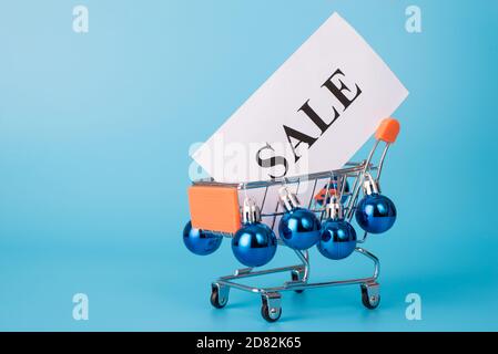 Shopping prima del concetto di Natale. Primo piano foto a lunghezza intera di divertente carrello carrello trolley di scherzo decorato con palle di albero giocattolo che trasportano banner con la vendita di testo Foto Stock