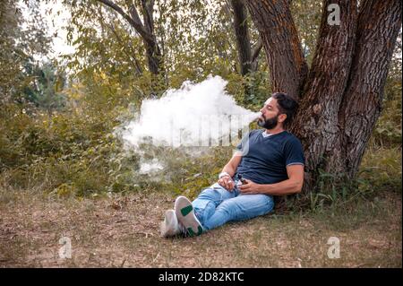 Uomo elegante che gode di un dispositivo elettronico di fumo in una foresta di pulizia. Attività di vaporizzazione. Foto Stock