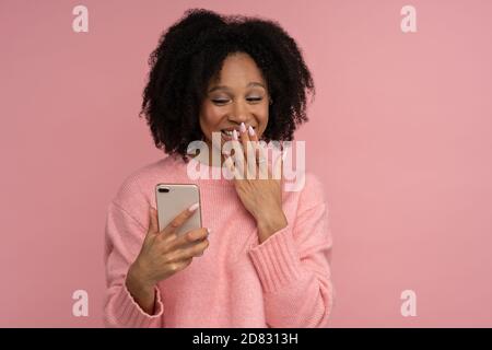 Sorridente donna millennial con pelle scura che guarda il telefono cellulare in shock, coprendo la sua bocca aperta con la mano, leggendo il messaggio del ragazzo, confus Foto Stock