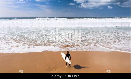 Vista dall'alto di un uomo surfista in muta con una tavola da surf in piedi vicino all'oceano sulla spiaggia sabbiosa.Pronto per il surf.Vacation hobby. Oceano Atlantico, Portogallo Foto Stock