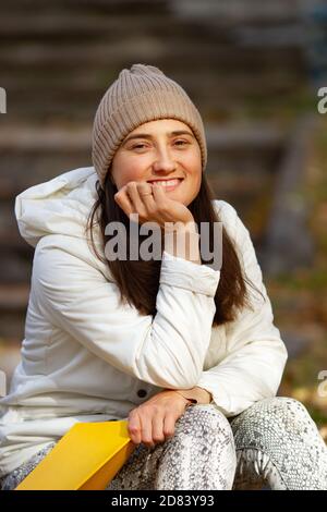 Felice bella donna brunetta seduta nel parco leggendo il libro in una bella giornata di sole autunno. Umore meditativo. Foto Stock