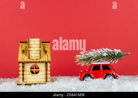 Piccola auto giocattolo rossa che trasporta l'albero di spruse su una strada di neve vicino al hous di legno. Biglietto d'auguri natalizio Foto Stock