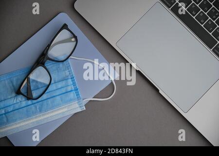 Flatlay di computer portatile, maschera e occhiali su una scrivania grigia. Lavorare da casa. Virus e autoisolamento Foto Stock