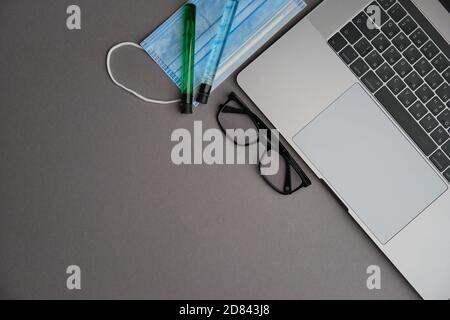 Flatlay di computer portatile, tubi medici, maschera e occhiali su una scrivania grigia. Lavorare da casa. Virus e autoisolamento Foto Stock