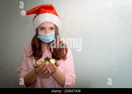 Una ragazza adolescente si alza a casa indossando un cappello di Santa e una maschera medica, tenendo i giocattoli d'oro dell'albero di Natale nelle sue palme Foto Stock