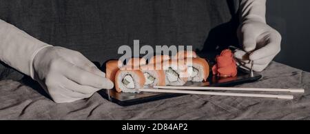 Le mani in guanti di gomma servono il sushi su un supporto nero. Menu sushi. Foto Stock