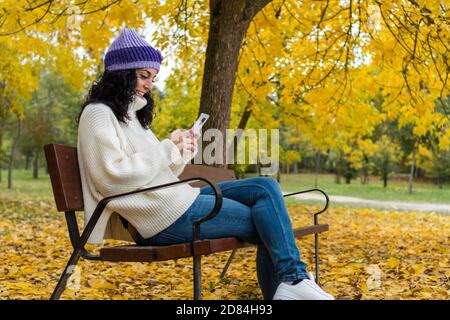 giovane donna con capelli neri ricci e caldo in maglione e cappello di lana seduto su una panchina di legno in un parco in autunno con foglie degli alberi che cadono dentro t Foto Stock