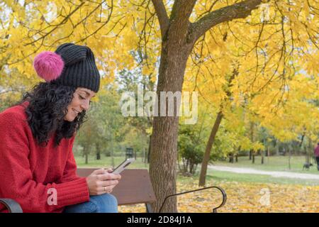 giovane donna con capelli ricci neri che indossa un maglione e. cappello di lana seduto su una panca di legno in un parco in autunno con foglie degli alberi che cadono nel Foto Stock