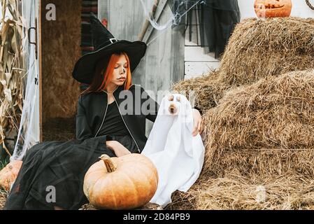 una ragazza in costume da strega con un cane dentro un costume fantasma che si diverte sul portico di a. Casa decorata per celebrare una festa di Halloween Foto Stock