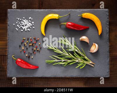 Erbe e spezie su sfondo ardesia. Vista dall'alto o piatta. Cibo di fondo e ingredienti alimentari - rosmarino, peperoni caldi, bulbi di aglio. Sale di casher e pepe Foto Stock