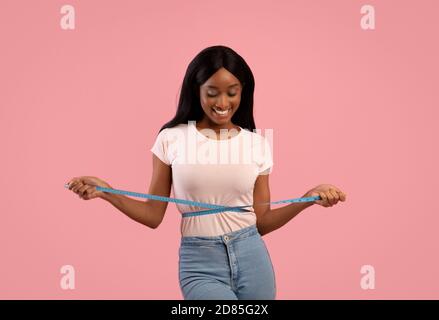 Perdita di peso e concetto di stile di vita sano. Donna nera sottile che misura la sua vita con nastro su sfondo rosa studio Foto Stock