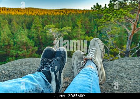 Immagine concettuale di due coppie di gambe in sneakers sullo sfondo della roccia forestale. Scarpe da ginnastica unisex casual e jeans in montagna. Coppia in cima alla montagna. Stile di vita. Foto Stock
