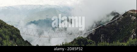 Vista grandangolare di un lungo ponte sospeso un abisso tra due colline nella nebbia con un splendida vista panoramica delle scogliere innevate e cl Foto Stock