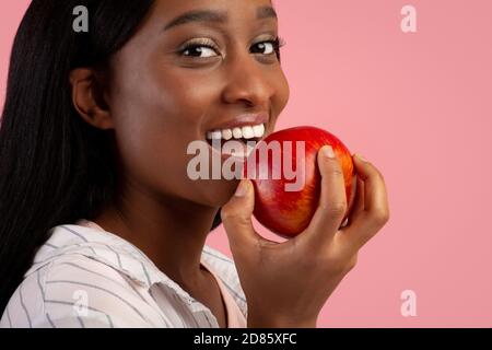 Concetto di denti sani. Bella donna nera con bel sorriso mordente mela rossa fresca su sfondo rosa studio Foto Stock