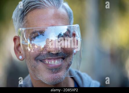 Deliziato cubano maschio in orecchini con scudo protettivo in plastica in piedi Sulla strada a Madrid durante l'epidemia di coronavirus e guardando fotocamera Foto Stock