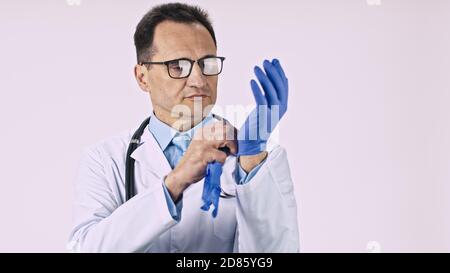 Serio medico maschile in camice di laboratorio e stetoscopio mette su guanti di protezione in lattice Foto Stock