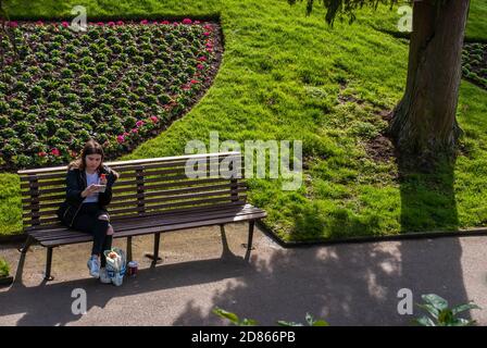 Guildford, Regno Unito - 05 maggio 2019 : UNA giovane donna seduta a leggere un messaggio di testo sul telefono cellulare su una panca di legno nel verde del parco. Nessuna messa a fuoco, speci Foto Stock