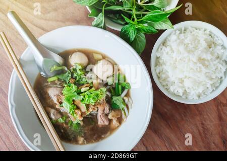 Zuppa di noodle di manzo brasata con palla di maiale in ciotola bianca e riso al vapore, foglie di basilico su tavolo di legno. Stile tailandese. Vista dall'alto. Disposizione piatta. Mettere a fuoco su noodle. Foto Stock