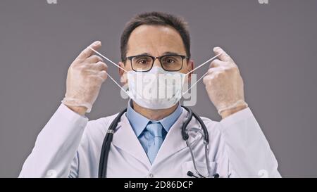 Bel tecnico di laboratorio in uniforme medica toglie la maschera protettiva e sorrisi Foto Stock