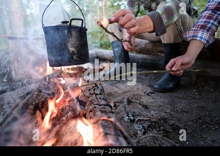 Due amici frittura dolce marshmallows su un fuoco con un bollitore sulla fiamma, in una giornata estiva, in un campeggio, nella foresta. Foto Stock