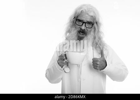 Studio shot di felice senior uomo barbuto medico sorridere mentre dando pollice in alto e tenendo premuto tazza da caffè Foto Stock