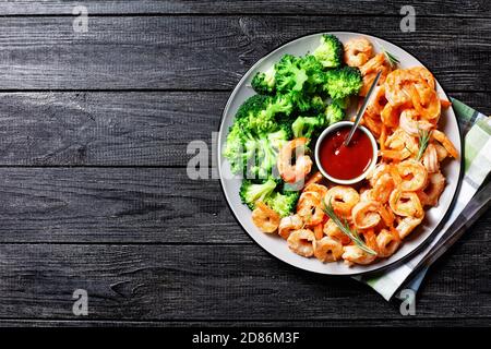 Gamberi o gamberetti al burro d'aglio disgelati e fritti serviti con broccoli al vapore, ketchup e lime su un piatto con rametti di rosmarino e posate su una d Foto Stock