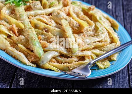 Fagioli di cera a basso contenuto calorico alla francese d'oro con aglio e prezzemolo ricoperti di pangrattato e fritti su una padella, serviti su un piatto blu con forchetta al buio Foto Stock