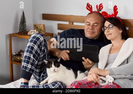 Felice coppia in pigiama di natale sdraiati sul letto e utilizzando tablet per videochiamare il giorno di natale per salutare la loro famiglia. Messa a fuoco morbida Foto Stock