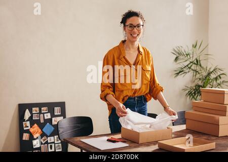 Giovane donna che guarda la macchina fotografica e sorride all'ufficio di affari di trasporto di goccia a casa. Sorridente in linea il proprietario di affari del deposito che impacchettano le scatole del pacco per deliv Foto Stock